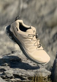 Lys beige treningstøvel med teksturert overdel, snøring og dempende såle, designet for utendørs aktiviteter.