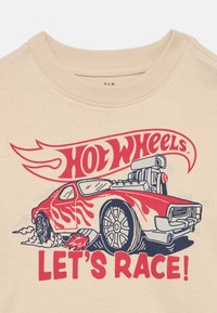 Μπεζ μπλουζάκι με γραφικό καρτούν αυτοκινήτου. Το αυτοκίνητο είναι κόκκινο με φλόγες, λεπτομερείς τροχούς και κείμενο που λέει «HOT WHEELS LET'S RACE!» σε κόκκινο χρώμα.