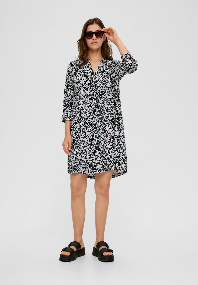 Schwarz-weiß gemustertes Kleid mit V-Ausschnitt und drei Viertelärmeln, mit asymmetrischem Saum und lockerer Passform. Getragen mit Plateausandalen.