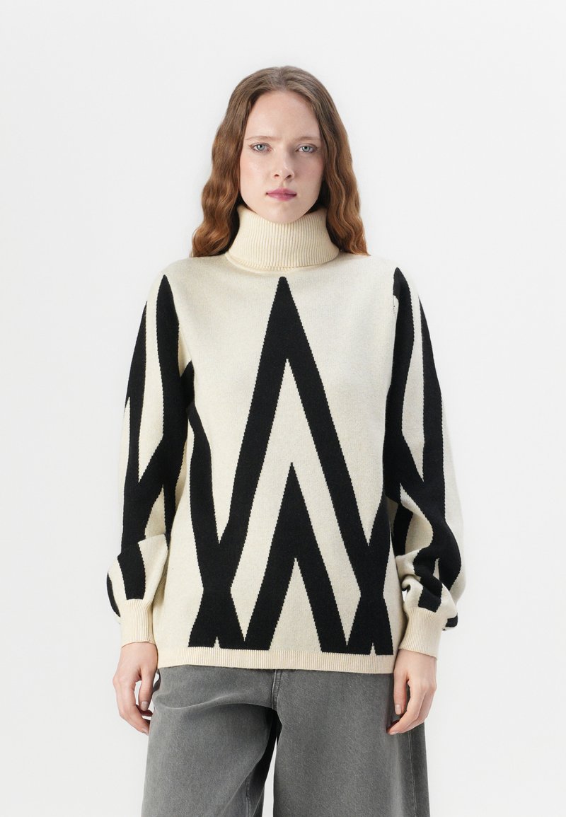 Pārmērīgs turtlenecks džemperis krēmkrāsā ar izteiksmīgu melnu zīmulīšu rakstu. Mīksta adīšanas tekstūra, nokritušie pleci un ribotas aproces.