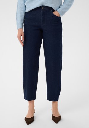 Kvinde iført mørkeblå højtsiddende jeans med lige ben, hænderne i lommerne, lyseblå sweater og brune spidse sko.