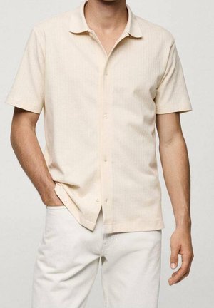 Homme portant une chemise beige à manches courtes boutonnée et un pantalon blanc, la main gauche dans la poche et le bras droit détendu le long du corps.