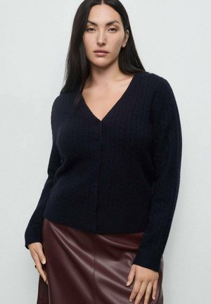 Cardigan de punto trenzado en color navy con escote en V y cierre de botones, con mangas largas y puños acanalados, acompañado de una falda de piel marrón.
