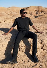 Homme en tenue noire et lunettes de soleil assis sur un sol rocheux dans un paysage désertique ensoleillé avec des collines beiges en arrière-plan.