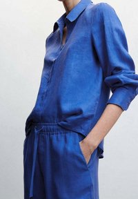 Chemise en lin bleue avec col, manches longues et coupe décontractée. Portée avec un pantalon bleu assorti à cordon de serrage. Texture lisse.