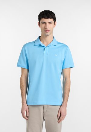 Mladić u svijetloplavoj polo majici kratkih rukava i bež hlačama, stoji ispred obične bijele pozadine.