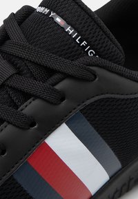 Chaussure de sport noire avec une tige en mesh, des lacets ronds et une semelle en caoutchouc. Détail distinctif avec une bande rouge, blanche et bleue sur le côté.