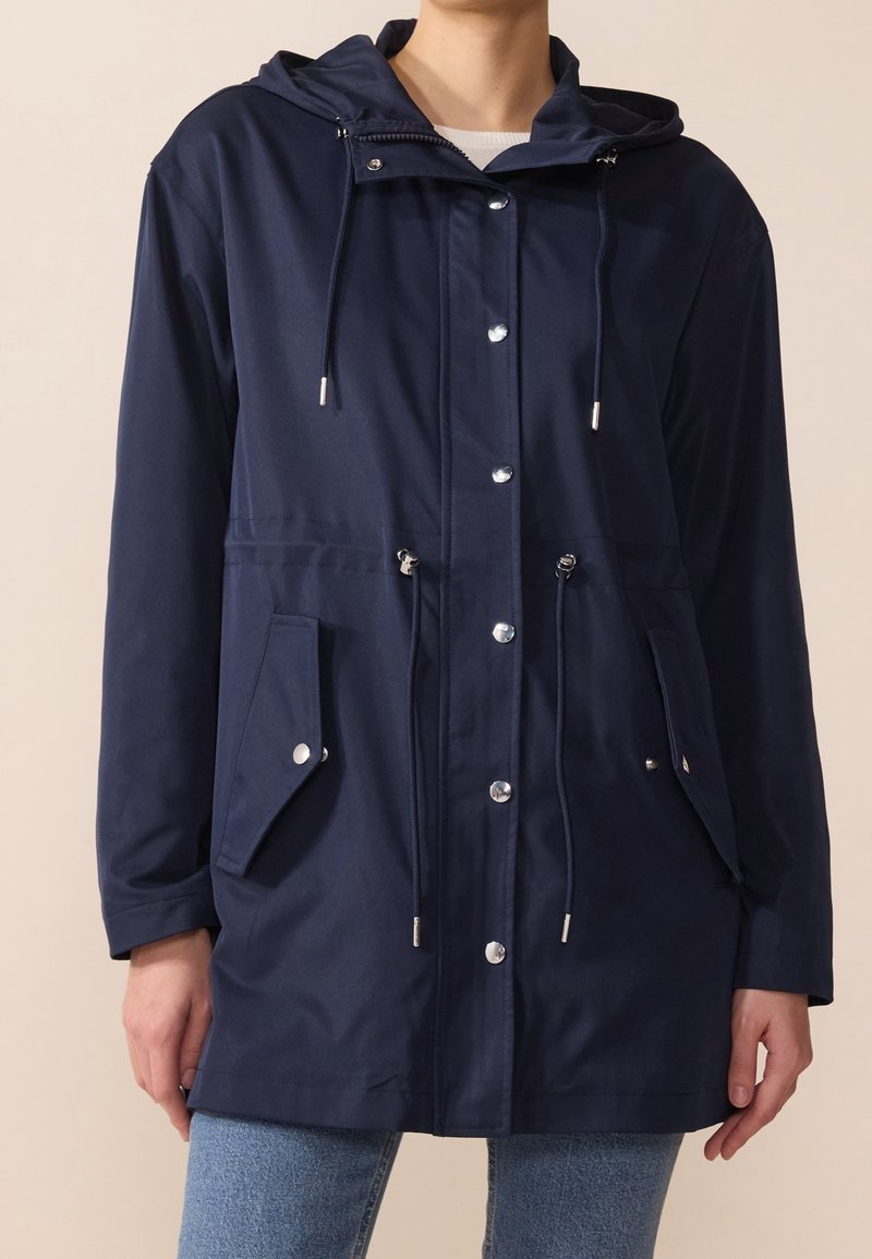 Marinblå vattentät jacka med huva, dragsko i midjan, tryckknappar och framfickor. Tillverkad av ett mjukt och lätt tyg.