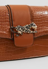 Ruskea nahkalaukku krokotiilikuviolla. Siinä on kultainen ja musta leopardilukko, sileä pinta ja jämäkkä muotoilu.