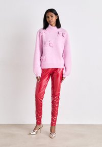 Růžový pletený svetr s mašlemi, spárovaný se světlými červenými latexovými kalhotami a stříbrnými lodičkami se špičatou špičkou. Hladká textura a živé barvy.