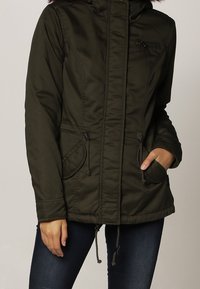 Kobieta ubrana w oliwkową ocieplaną kurtkę z zamkiem z przodu, z regulowanym sznurkiem w pasie i na dole kurtki, trzymająca rękę w prawej kieszeni.