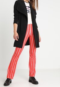 Černý dlouhý kabát přes světlou košili s grafickým potiskem, doplněný červenými proužkovanými kalhotami a černými šněrovacími botami.