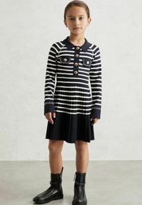 Gestreiftes Kleid in Marineblau und Weiß mit langen Ärmeln, Kragen, zwei Fronttaschen und plissiertem Rock. Kombiniert mit schwarzen Knöchelstiefeln mit Reißverschlüssen.