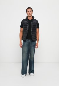 Vesta neagră cu umplutură, cu fermoar în față, purtată peste un tricou negru. Asociată cu blugi lejeri din denim albastru și adidași albi.