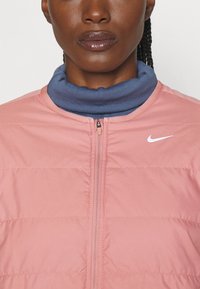 Veste doudoune rose clair avec une fermeture éclair sur le devant, dotée d'un col montant bleu. Texture brossée et logo Nike blanc sur la poitrine.