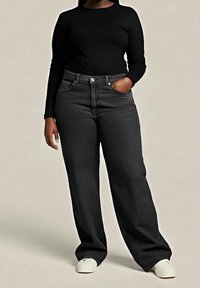 Personne portant un haut noir à manches longues, un jean noir ample taille haute, des chaussures blanches, avec une main dans la poche sur un fond uni.