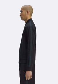 Giacca sportiva nera con zip, caratterizzata da una striscia verde a motivo sulle maniche e tasche laterali, indossata con pantaloni coordinati.