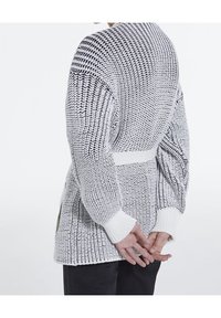 Person som bär en texturerad vit stickad tröja med svarta detaljer, med händerna knäppta bakom ryggen, tillsammans med svarta byxor.