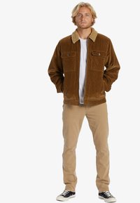 Nuori mies seisoo kädet taskuissa, päällään ruskea corduroy-takki, jossa on keinoturkiskaulus, valkoinen paita, beige housut ja mustat lenkkarit.