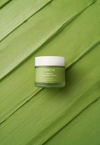 Tirtir Matcha rauhoittavan voiteen purkki, jossa valkoinen kansi, teksturoidulla vihreällä taustalla, joka vastaa voiteen väriä ja rakennetta.