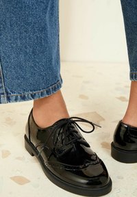 Pantofi oxford din piele lăcuită neagră, cu vârf rotunjit, model cu șireturi și detalii perforate, asortați cu blugi bleu cu mansete.