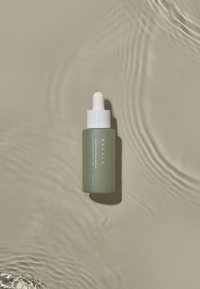 Bottiglia contagocce in vetro verde con tappo bianco, caratterizzata da un testo semplice e un design elegante, posizionata su una superficie beige chiaro con ondulazioni.
