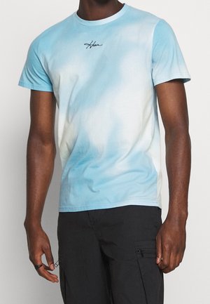 Homme portant un t-shirt manches courtes tie-dye bleu clair et blanc avec un petit texte noir sur la poitrine et un pantalon cargo noir.