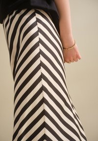 Kobieta ubrana w czarno-białą spódnicę w jodełkę i czarną bluzkę, z złotą bransoletką i pierścionkiem na prawej ręce.