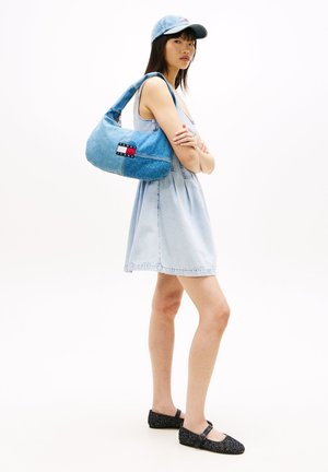 Kvinde iført lyseblå cowboykjole og kasket, bærende en blå Tommy Jeans skuldertaske, stående med armene krydsede og iført sorte flade sko.