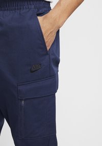 Námornické cargo kalhoty s bočními kapsami, elastickým pasem a černým logem Nike. Texturovaná tkanina s hladkým povrchem a viditelným detailem na zipu.