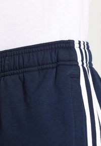 Ceinture élastique bleu marine sur un pantalon de survêtement avec trois bandes blanches verticales et une partie d'une chemise blanche rentrée.