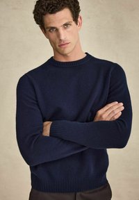 Navyblauwe gebreide trui met een ronde halslijn, geribde manchetten en zoom, gemaakt van een zachte stof, aansluitende stijl, gedragen door een model met gekruiste armen.