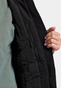 Giacca nera con una consistenza liscia e trapuntata; presenta una tasca con zip e una chiusura a scatto; fodera del tessuto visibile in un verde morbido.