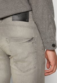Jeans in denim grigio con texture leggera, caratterizzati da una tasca posteriore cucita in un design curvo e un'etichetta in pelle nera sul passante della vita.