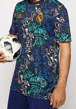 Homme tenant un ballon de football Adidas, portant un maillot noir du Real Madrid avec des motifs et logos style graffiti en bleu, blanc et sarcelle.