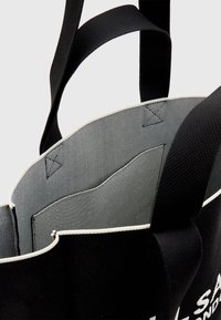 Sac cabas noir avec larges poignées, doublure intérieure gris clair et poche intérieure sur un fond neutre.