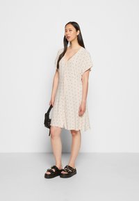 Vestido bege claro com bolinhas pretas, mangas curtas, decote em V, parte da frente com botões e uma barra rodada, usado com sandálias de plataforma pretas.