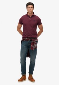 Mørkerød polo shirt med opretstående krave, rutet skjorte bundet om livet, smalle jeans og brune snørebåndsstøvler. Glat tekstur, afslappet design.