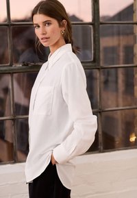 Femme portant une chemise blanche boutonnée avec une poche avant, debout de profil avec une main dans la poche d’un pantalon noir, devant une grande fenêtre.