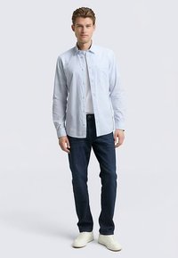 Camicia azzurro chiaro abbottonata sopra una t-shirt bianca, abbinata a jeans blu scuro e sneakers bianche, in piedi su uno sfondo semplice.