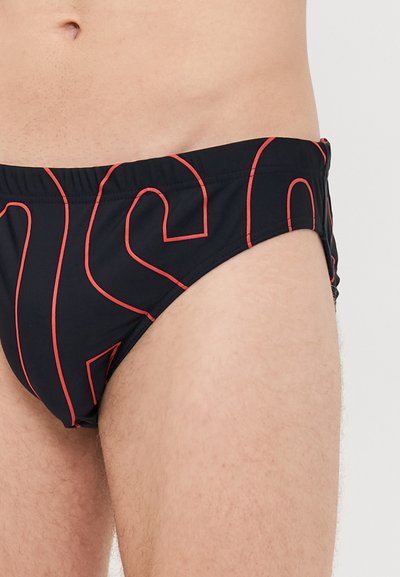 Torso masculin et parties supérieures des jambes portant un slip de bain noir avec des lettres rouges sur un fond clair.