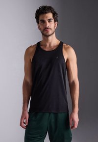 Svart atletiskt linne med en slät textur. Har rund halsringning och vida armhål, kombinerat med mörkgröna shorts. Diskret logodetalj.