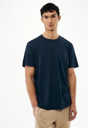 Junger Mann mit kurzen, lockigen Haaren, der ein schlichtes, marineblaues T-Shirt mit kurzen Ärmeln und beige Hosen trägt, vor einem schlichten weißen Hintergrund steht.