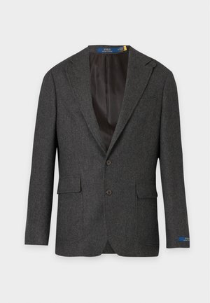 Homme portant un blazer gris foncé, une chemise blanche boutonnée, un pantalon marron et des chaussures habillées noires, debout avec une main dans la poche.