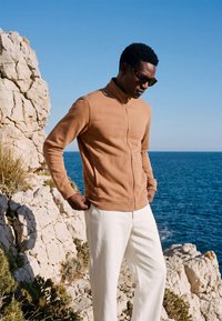 Homme portant des lunettes de soleil, une veste zippée beige et un pantalon blanc, debout sur des falaises rocheuses au bord de la mer sous un ciel bleu clair.