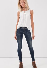 Vit ärmlös topp med en texturerad frampanel, kombinerad med mörkblå skinny jeans och bruna ankelboots. Enkel, ren design.