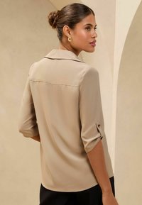 Blusa beige con textura suave, mangas tres cuartos y puños enrollados que presentan detalles de botones dorados. Cuello clásico y línea de costura sutil.
