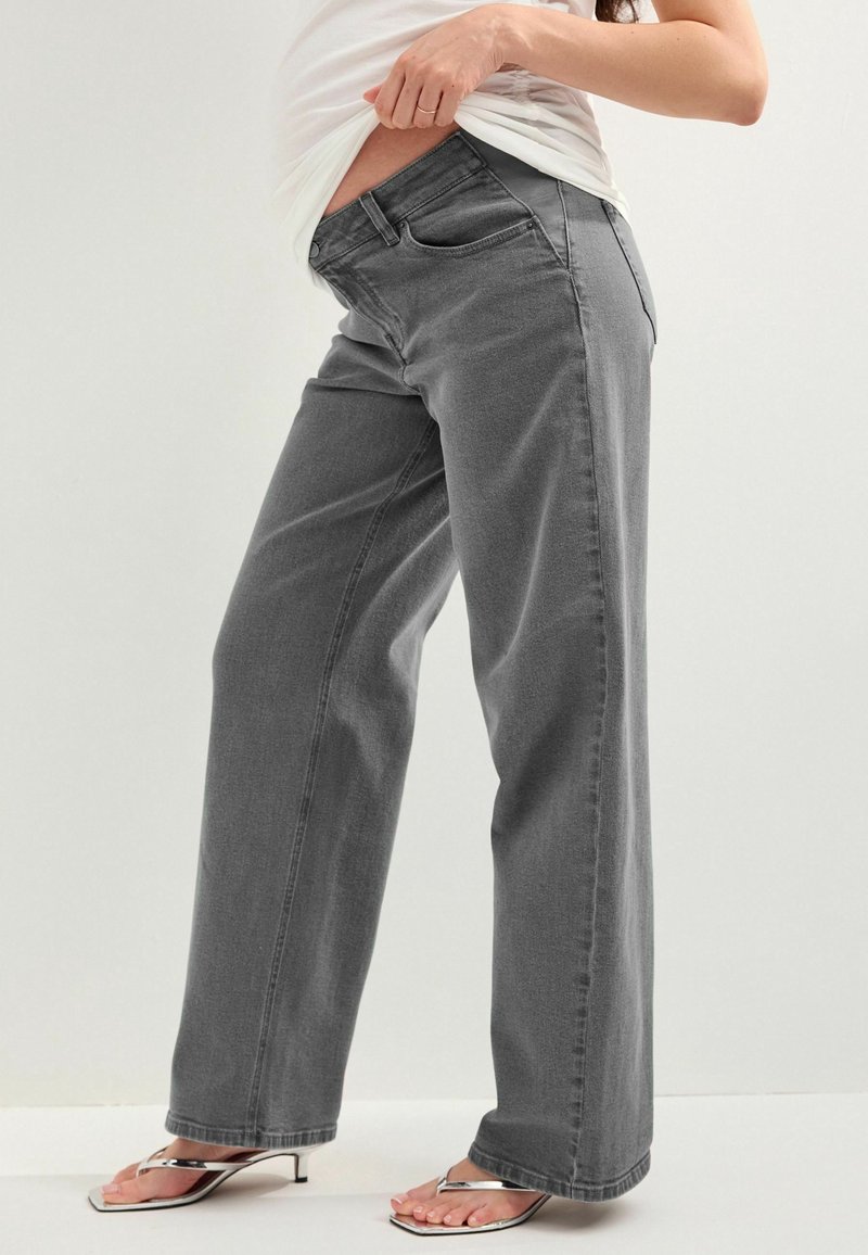 Personne portant un jean de maternité gris ample, levant une chemise blanche pour montrer son ventre de femme enceinte, associée à des sandales à talons argentées.