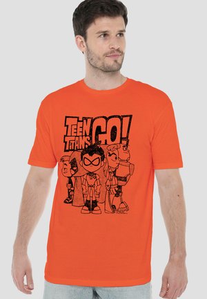 Mann trägt ein helloranges T-Shirt mit schwarzen Teen Titans Go! Cartoonfiguren und Text vorne, steht vor einfarbigem Hintergrund.