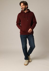 Bordeaux hoodie met een voorzak en koordjes, gecombineerd met blauwe jeans. Model draagt beige sneakers op een neutrale achtergrond.